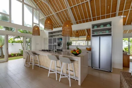 Spacious modern Balinese-style kitchen with high wooden ceiling, rattan pendant lights, white island bar, fresh fruits, and tropical garden views through large windows. - Bali Villas